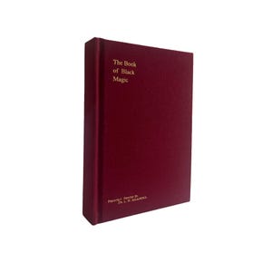 May include: A burgundy book titled "The Book of Black Magic" with gold lettering. The book has a textured cover and is standing upright. The bottom of the book reads "Privately Printed By Dr. L. W. de Laurence."