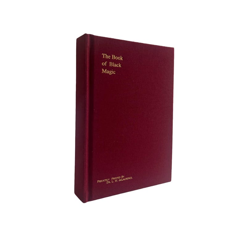 May include: A burgundy book titled "The Book of Black Magic" with gold lettering. The book has a textured cover and is standing upright. The bottom of the book reads "Privately Printed By Dr. L. W. de Laurence."