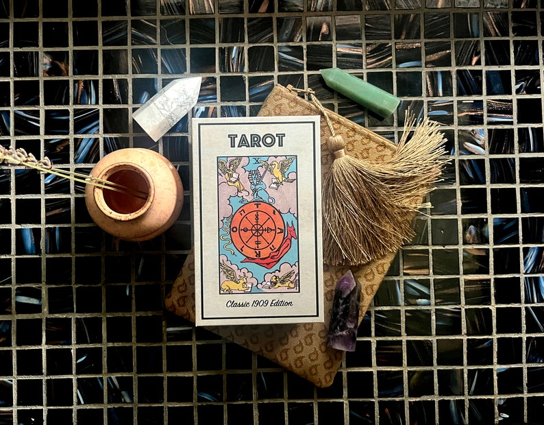Puede incluir: Una caja de baraja de tarot con un borla dorada y un cristal verde. La caja est&aacute; abierta y muestra la carta de la Rueda de la Fortuna de una baraja de tarot cl&aacute;sica de 1909.