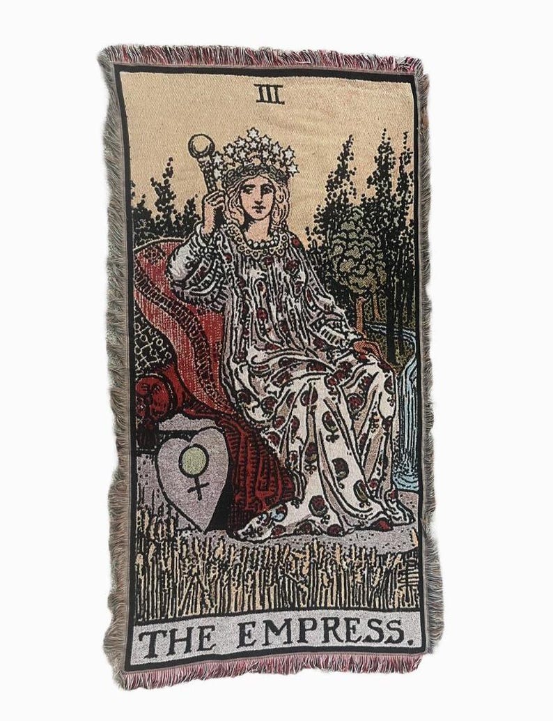 May include: A tapestry featuring the Empress card from a tarot deck. The Empress is depicted sitting on a throne with a crown on her head and a scepter in her hand. The card is surrounded by a border of flowers and leaves. The text "THE EMPRESS" is printed at the bottom of the card.