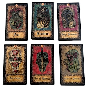 May include: A set of ten tarot cards with black borders and ornate gold frames. The cards feature illustrations of skulls and other imagery. The card names are printed in black at the bottom of each card. The card names are: Fool, Magician, High Priestess, Two of Cups, Ace of Swords, Queen of Pentacles.