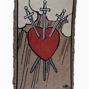 May include: A tapestry depicting the Three of Swords tarot card. The card features a red heart pierced by three swords, symbolizing heartbreak and sorrow. The background is a beige color with clouds and a brown border.