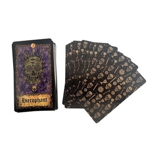 May include: A tarot deck with a black background and gold accents. The card shown is the Hierophant, which features a skull with multiple eyes and a purple background. The back of the cards features a repeating pattern of skulls and keys.