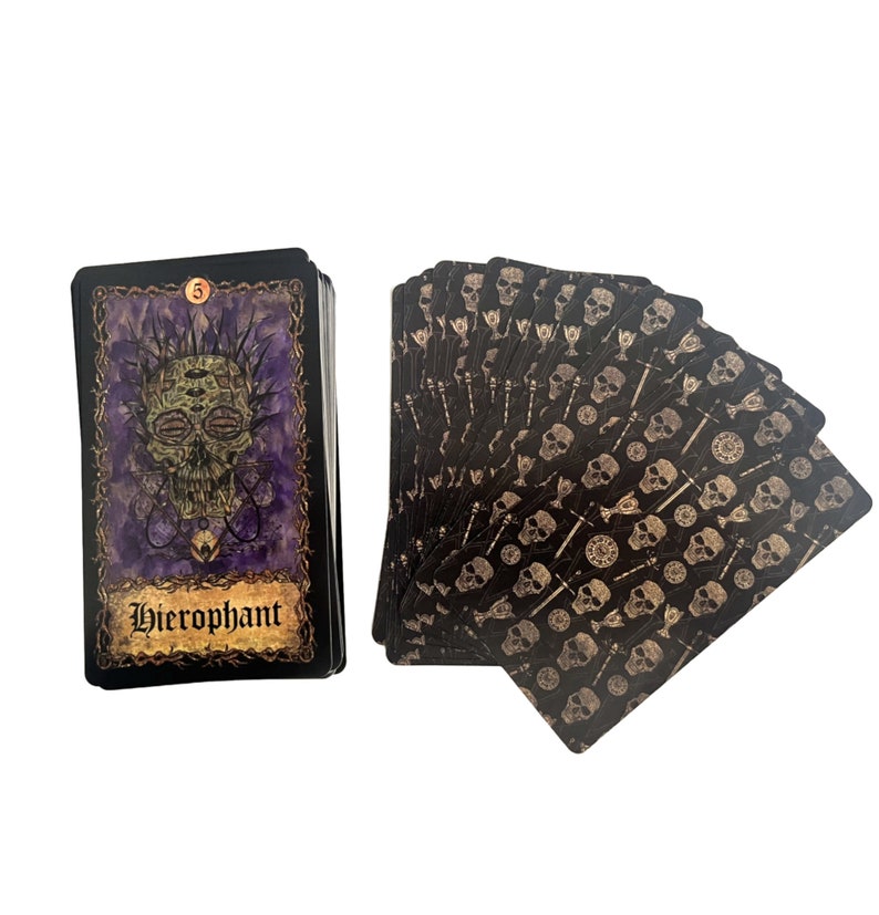 May include: A tarot deck with a black background and gold accents. The card shown is the Hierophant, which features a skull with multiple eyes and a purple background. The back of the cards features a repeating pattern of skulls and keys.