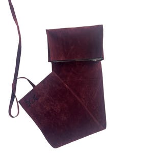 Puede incluir: Una bolsa de ante burdeos con una correa larga y un cierre de solapa. La bolsa tiene un s&iacute;mbolo de triple luna bordado en la parte delantera.