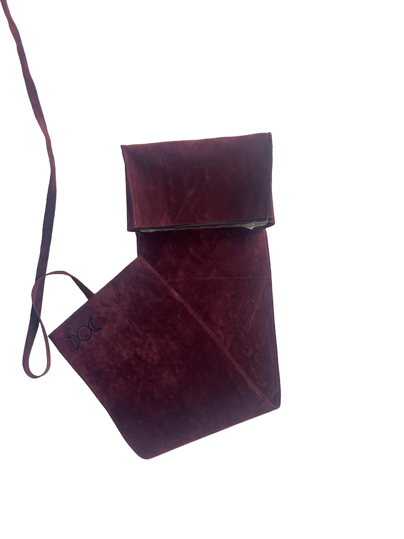 Puede incluir: Una bolsa de ante burdeos con una correa larga y un cierre de solapa. La bolsa tiene un s&iacute;mbolo de triple luna bordado en la parte delantera.