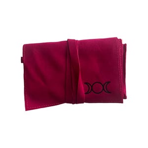 May include: A pink velvet pouch with a black triple moon symbol embroidered on the front. The pouch has a flap closure and a strap.
