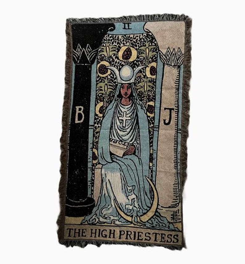 May include: A tapestry featuring the High Priestess card from the tarot deck. The card depicts a woman wearing a blue robe and a crown with a crescent moon. She is seated between two pillars, one black and one white, with the letters B and J on them. The card is framed with a brown border and has a fringe.