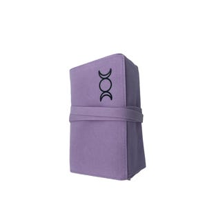 Puede incluir: Una bolsa de fieltro lavanda con un s&iacute;mbolo de triple luna negro. La bolsa est&aacute; enrollada y atada con una correa.