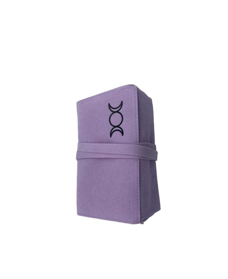 Puede incluir: Una bolsa de fieltro lavanda con un s&iacute;mbolo de triple luna negro. La bolsa est&aacute; enrollada y atada con una correa.