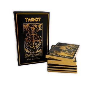 以下が含まれることがあります： 黒と金のタロットカードデッキ。「TAROT」と「WHEEL of FORTUNE」の文字が箱に書かれています。カードは金色の縁取りで、一番上のカードには「THE FOOL」と書かれています。