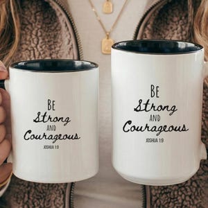 May include: Two white ceramic mugs with black interiors and handles. Each mug features the black printed text "BE Strong AND Courageous" followed by "JOSHUA 1:9" in a mix of serif and script fonts. The mugs are held side by side.