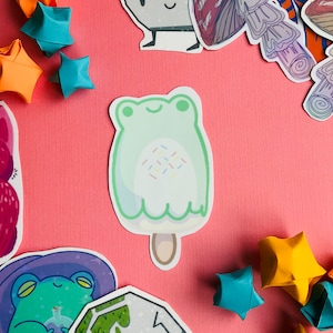 May include: A green frog-shaped popsicle with white frosting and colorful sprinkles. The popsicle is on a pink background.