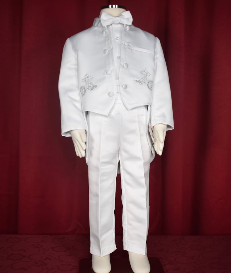 May include: A white suit for a young boy, featuring a jacket with a tailcoat, a vest, and matching pants. The jacket has a stand-up collar, a bow tie, and silver embroidery of crosses on the front.