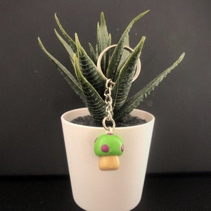 Op de afbeelding: Een groene paddenstoel sleutelhanger met paarse stippen en een beige steel. De sleutelhanger hangt aan een zilveren ketting en ring.