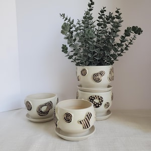 May include: A set of four off-white ceramic plant pots with matching saucers. Each pot features unique brown abstract designs. One pot is stacked on top of two others, with a fourth pot in front. The stacked pots hold a green leafy plant.