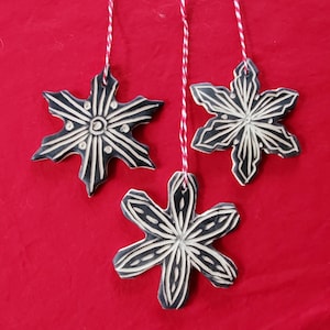 Può includere: Tre ornamenti a forma di fiocco di neve in bianco e nero appesi su uno sfondo rosso. Ogni ornamento è realizzato in ceramica e presenta un design intagliato unico. Gli ornamenti sono sospesi da uno spago rosso e bianco.