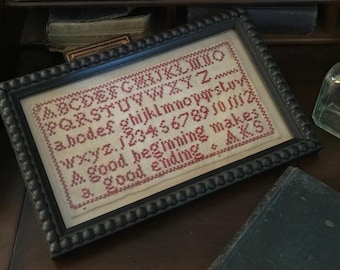 Könnte beinhalten: Ein gerahmter Kreuzstich-Sampler mit roter Stickerei auf weißem Stoff. Der Sampler enthält das Alphabet, Zahlen und den Spruch "Ein guter Anfang macht ein gutes Ende."