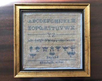 May include: Framed antique sampler with blue cross-stitch lettering and crown motifs. The sampler includes the alphabet, numbers, and the name "Elizabeth Arker" with the word "England" below. The frame is gold-colored.