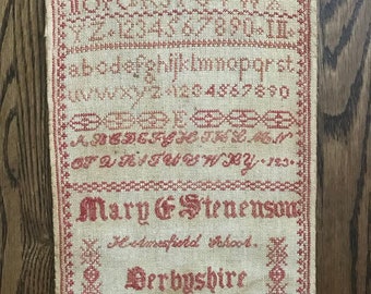 May include: A vintage sampler with red cross-stitch embroidery on a beige linen background. The sampler features the alphabet, numbers, and the name "Mary G. Stevenson," along with "Holmesfield School" and "Derbyshire 1876."