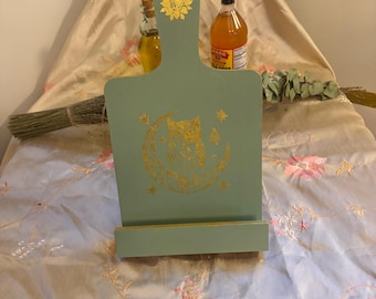 Könnte beinhalten: Ein salbeigrüner Kochbuchständer aus Holz mit einem goldenen Sonnenblumenmotiv an der Oberseite und einer goldenen Grafik einer Eule auf einer Mondsichel in der Mitte. Der Ständer verfügt über eine rechteckige Ablagefläche für Bücher oder Tablets.