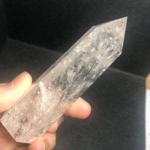 May include: A clear quartz crystal point with a faceted, tapered shape. The crystal is held in a hand and has internal inclusions and a slightly cloudy appearance. The crystal is a light, neutral color.