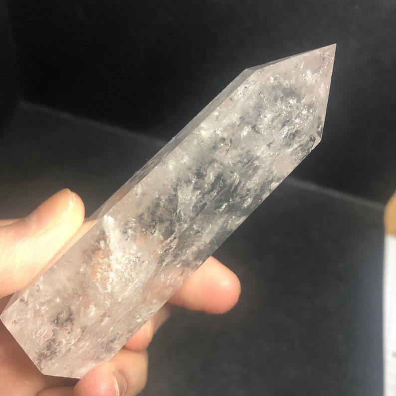 May include: A clear quartz crystal point with a faceted, tapered shape. The crystal is held in a hand and has internal inclusions and a slightly cloudy appearance. The crystal is a light, neutral color.