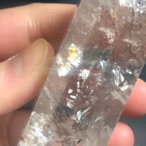 May include: A clear quartz crystal with internal fractures and iridescent rainbow reflections. The crystal is held in a hand, showcasing its polished surface and natural variations. The overall effect is a display of light and color.