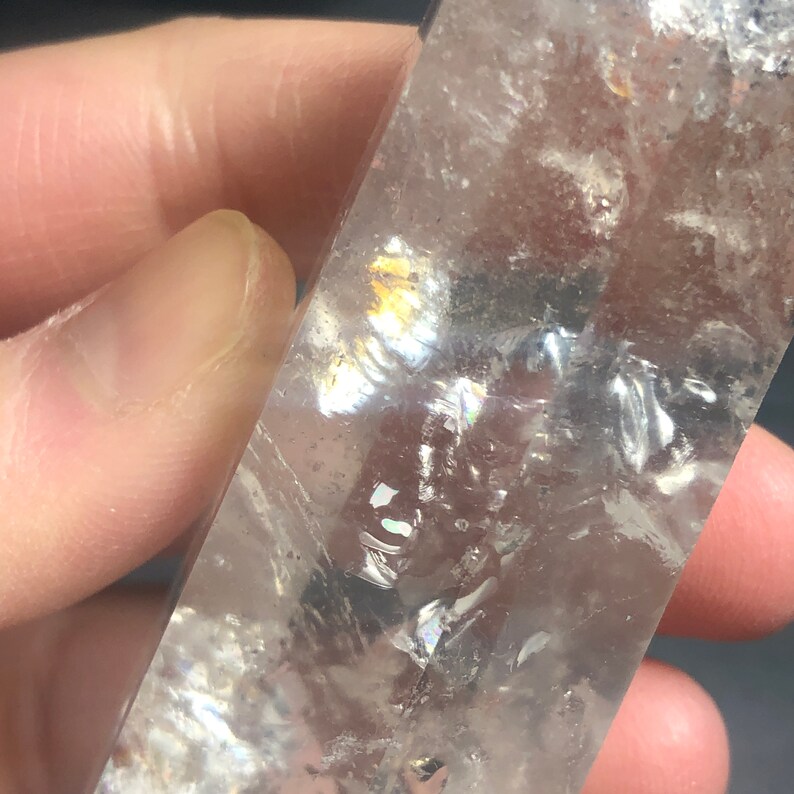 May include: A clear quartz crystal with internal fractures and iridescent rainbow reflections. The crystal is held in a hand, showcasing its polished surface and natural variations. The overall effect is a display of light and color.