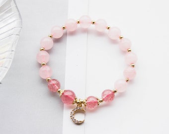 Könnte beinhalten: Ein rosa Perlenarmband mit einem goldenen Halbmond-Anhänger. Das Armband hat abwechselnd rosa und rot getönte Perlen, die durch kleine goldfarbene Perlen verbunden sind. Der Anhänger ist mit kleinen, klaren Kristallen besetzt.