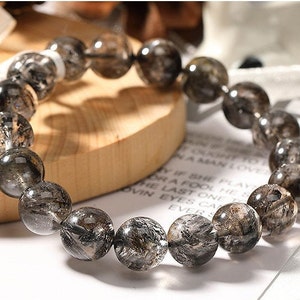 May include: A beaded bracelet featuring large, round, translucent beads with dark inclusions. The beads are strung together, forming a circular shape. The bracelet is displayed on a wooden surface, with text visible in the background.