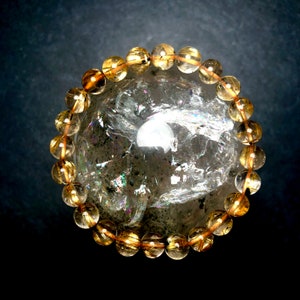 May include: A bracelet of golden-yellow, translucent beads strung on a thin, brown cord. The bracelet is resting on a large, clear, crystal geode with iridescent reflections. The background is a dark, gradient blue.
