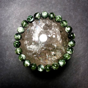 Puede incluir: Un brazalete de cuentas con piedras de serpentina verde oscuro y blanca. El brazalete es circular, con cada cuenta de aproximadamente 1 cm de diámetro. Las cuentas están dispuestas alrededor de una esfera de cristal transparente y texturizada, sobre un fondo gris oscuro.