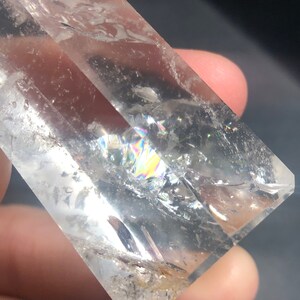 May include: A clear quartz crystal with internal fractures and rainbow iridescence. The crystal is held in a hand, showcasing its natural clarity and light refraction. The crystal's facets are visible, highlighting its crystalline structure.
