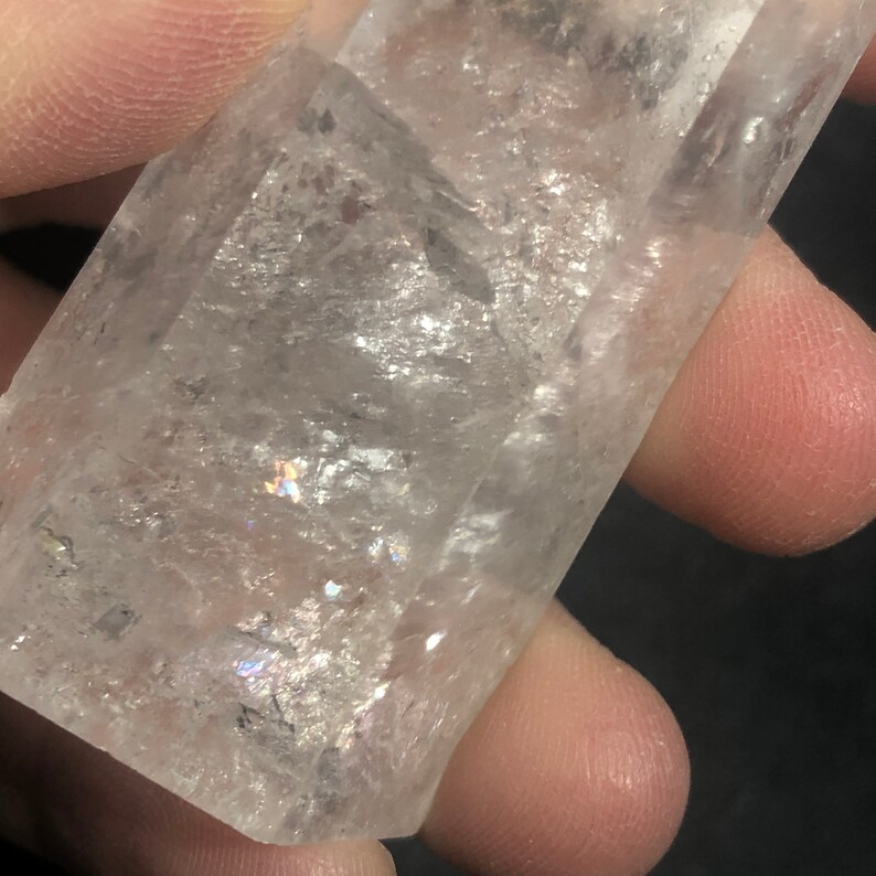 May include: A clear quartz crystal with a rectangular shape. The crystal has a slightly rough texture and displays internal rainbows. The crystal is held in a hand, with the fingers visible in the background. The crystal is a natural gemstone.