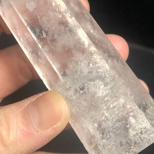 May include: A clear quartz crystal point with a rectangular shape. The crystal is held in a hand and has a textured surface with internal inclusions. The crystal is transparent and appears to be polished.