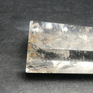 May include: A clear, rectangular crystal with visible internal fractures and inclusions. The crystal has a polished, smooth surface and a slightly cloudy appearance. The edges are sharp and well-defined, with some areas showing a hint of orange and brown coloration.