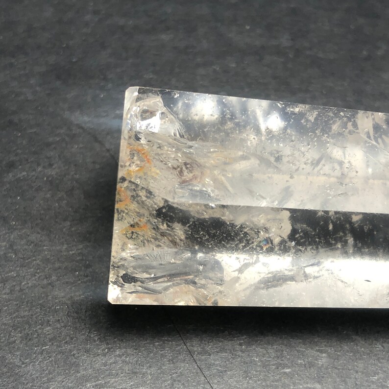 May include: A clear, rectangular crystal with visible internal fractures and inclusions. The crystal has a polished, smooth surface and a slightly cloudy appearance. The edges are sharp and well-defined, with some areas showing a hint of orange and brown coloration.