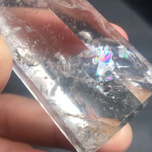 May include: A clear quartz crystal with internal fractures and rainbow iridescence. The crystal is rectangular and appears to be held by a person. The crystal is transparent, allowing light to pass through and create a prism effect.
