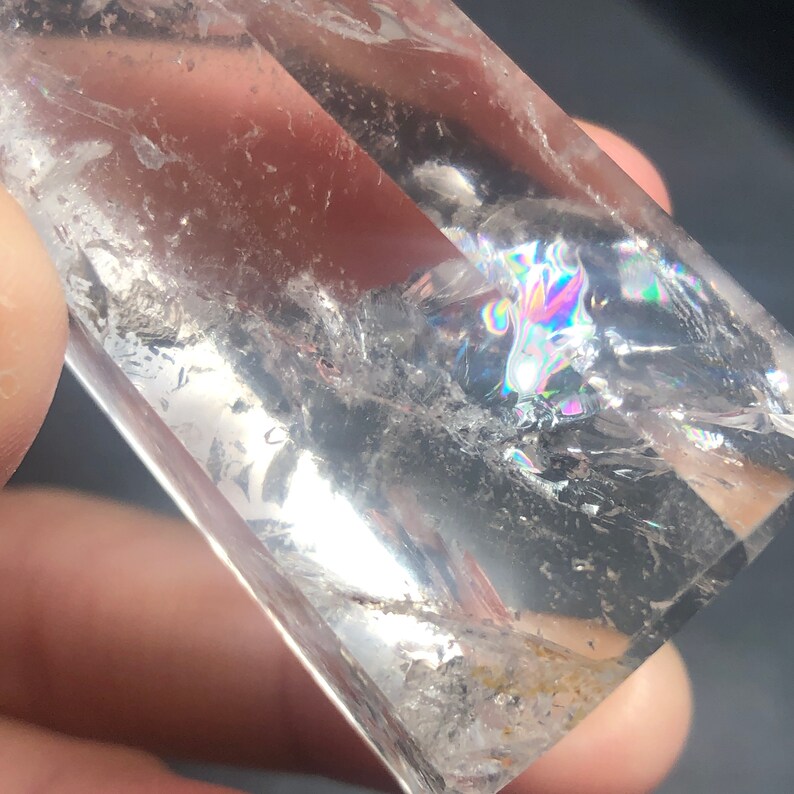 May include: A clear quartz crystal with internal fractures and rainbow iridescence. The crystal is rectangular and appears to be held by a person. The crystal is transparent, allowing light to pass through and create a prism effect.