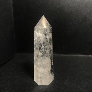 May include: A clear quartz crystal point with a hexagonal base and a pointed top. The crystal is transparent with internal inclusions, and it stands upright against a dark background. The crystal is approximately 5 inches tall.