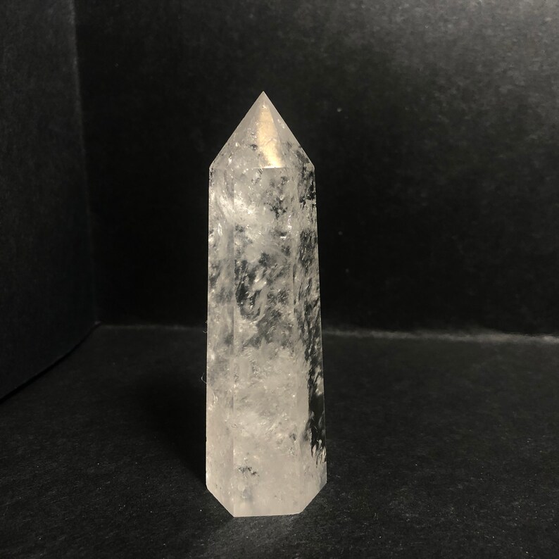 May include: A clear quartz crystal point with a hexagonal base and a pointed top. The crystal is transparent with internal inclusions, and it stands upright against a dark background. The crystal is approximately 5 inches tall.