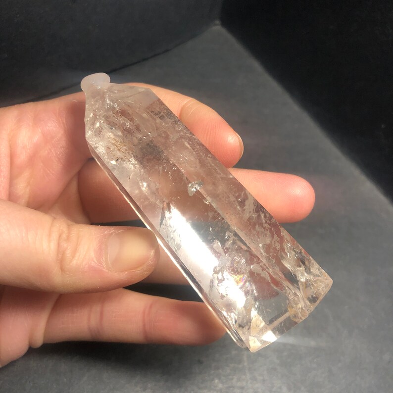 May include: A clear quartz crystal point with a polished, rectangular shape. The crystal is held in a hand, showcasing its natural clarity and subtle internal inclusions. The top of the crystal has a small, rounded tip.