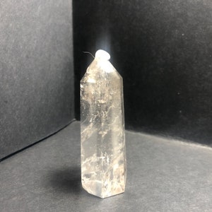 May include: A clear quartz crystal point with a hexagonal base and a tapered top. The crystal is transparent with internal fractures and inclusions, standing upright against a dark background. The crystal measures approximately 4 inches in height.