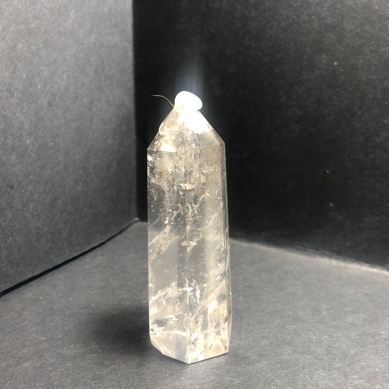 May include: A clear quartz crystal point with a hexagonal base and a tapered top. The crystal is transparent with internal fractures and inclusions, standing upright against a dark background. The crystal measures approximately 4 inches in height.