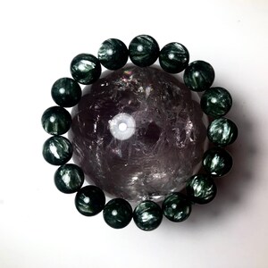 Puede incluir: Una pulsera de cuentas esféricas verde oscuro rodea una gran esfera de cristal translúcido, morado-gris. Las cuentas de la pulsera tienen un aspecto veteado, y la esfera de cristal tiene un interior texturizado y nublado. El fondo es blanco.