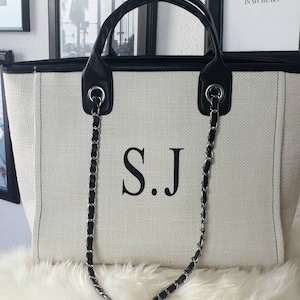 Può includere: Una borsa tote in tela bianca con finiture in pelle nera e una tracolla a catena nera. La borsa ha le iniziali "S.J." stampate in nero sul davanti.