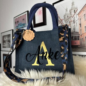 Könnte beinhalten: Eine marineblaue Tragetasche mit einem goldenen "A" und dem Namen "Anke" auf der Vorderseite bestickt. Die Tasche hat einen Riemen mit Leopardenmuster.