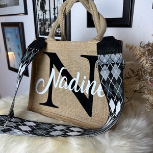 Könnte beinhalten: Eine beige Jute-Tragetasche mit einem schwarz-weißen, rautenförmigen Riemen. Die Tasche hat einen großen schwarzen Buchstaben "N" auf der Vorderseite mit dem Namen "Nadine" in weißen Buchstaben.