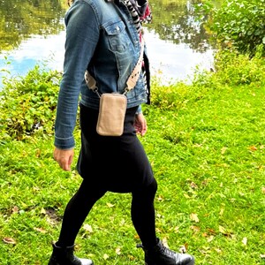 以下が含まれることがあります： デニムジャケット、黒いスカート、黒いタイツを着用した女性が草地のエリアを歩いている。彼女は、ストラップ付きの小さな薄い茶色のクロスボディバッグを持っている。